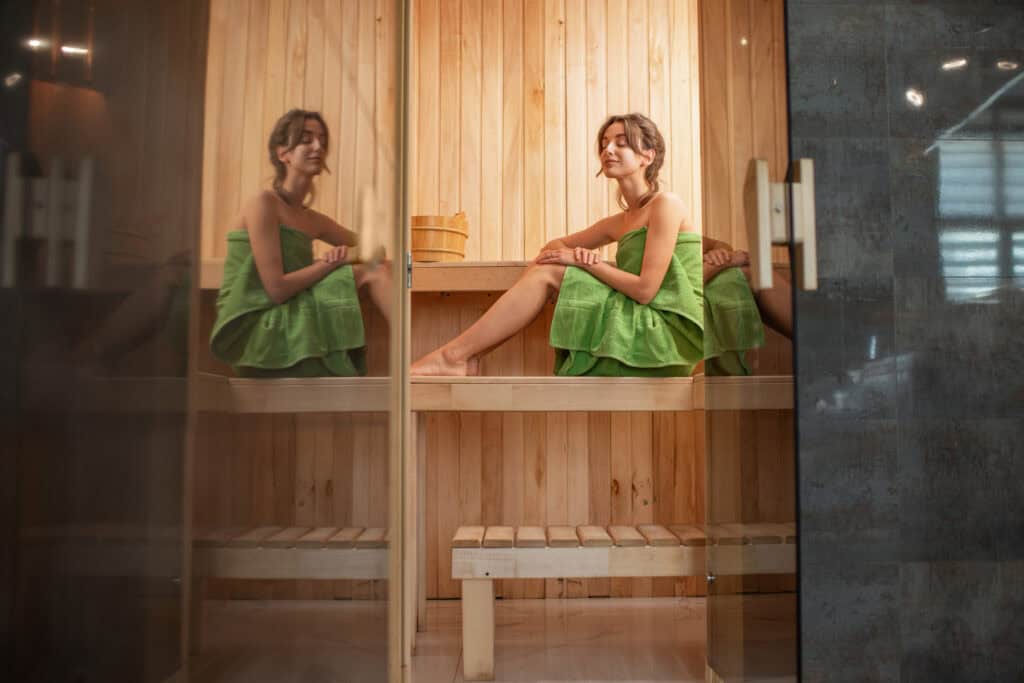 A woman wrapped in a green towel sits relaxed on a wooden bench inside an outdoor sauna, with her reflection visible on the glass wall in front of her.