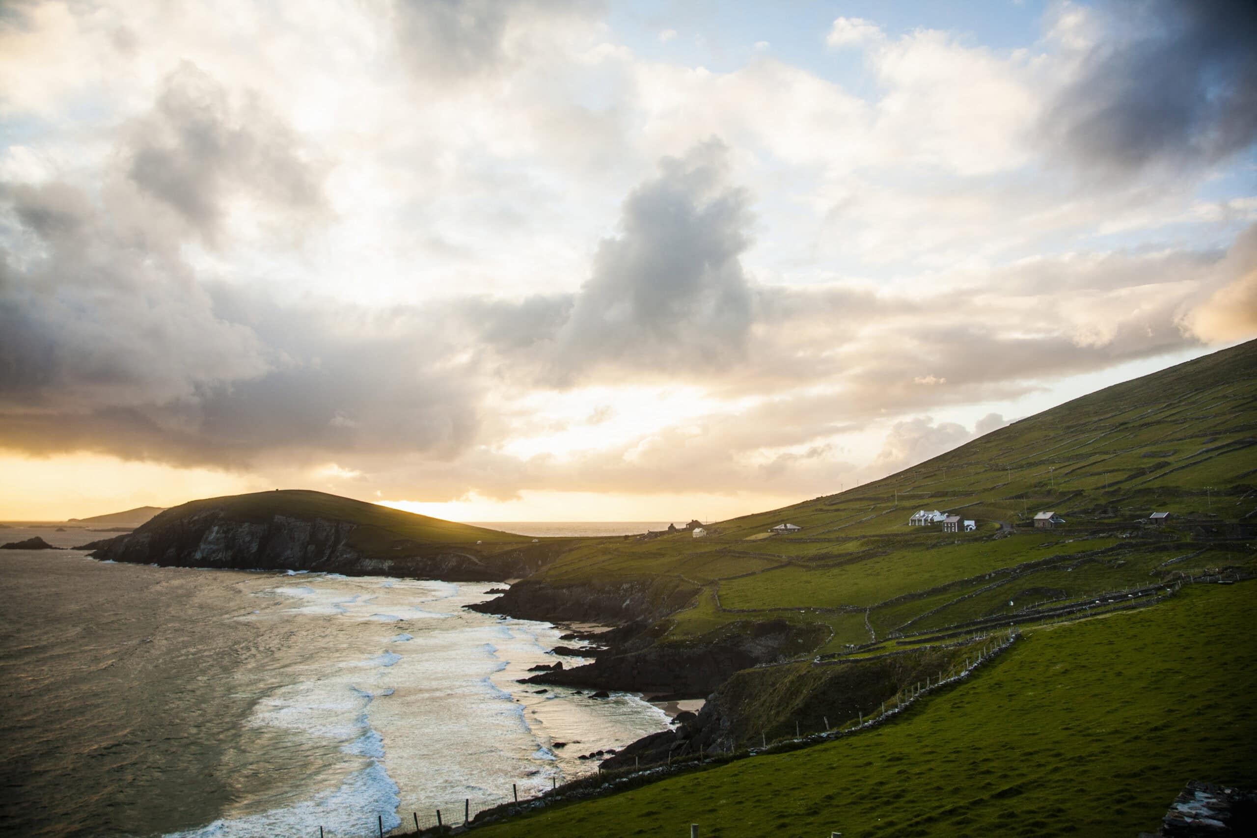 Sunset over a rugged coastline with rolling green hills, scattered houses, and waves crashing against the cliffs under a dramatic sky—an inviting spot for an outdoor sauna or cold plunge tub nestled amidst the breathtaking scenery.