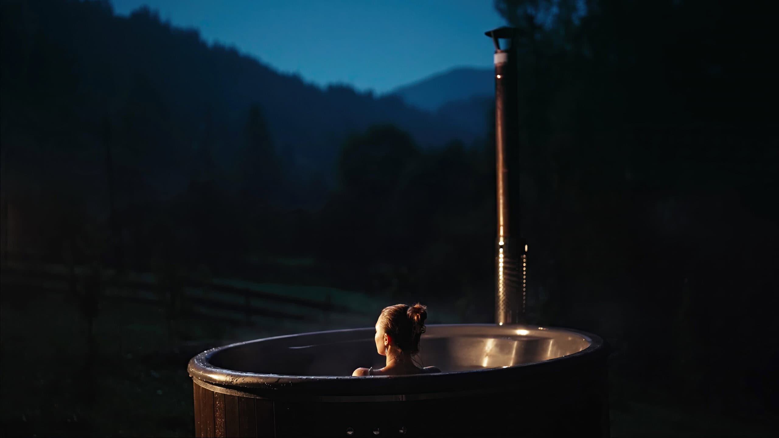 A person with hair in a bun relaxes alone in an outdoor hot tub at dusk, surrounded by dark trees and mountains under a deep blue sky, with a nearby barrel sauna adding to the serene, steamy atmosphere. Steam rises gently from the water.