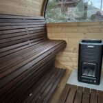 Wooden barrel sauna interior with curved bench seating, a Harvia sauna heater on the right, and a large window overlooking evergreen trees and a cabin outside.