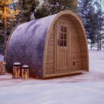 A small, arched wooden cabin and a cosy barrel sauna sit on a snow-covered decking in a winter forest. Two lanterns glow at the front with a stack of firewood nearby, while pine trees and snow create a peaceful backdrop.