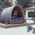 A small, modern arched garden sauna with glass doors sits on a wooden decking in a snowy landscape, surrounded by pine trees. A lit lantern decorates the decking, snow is gently falling, and a house is visible in the background.
