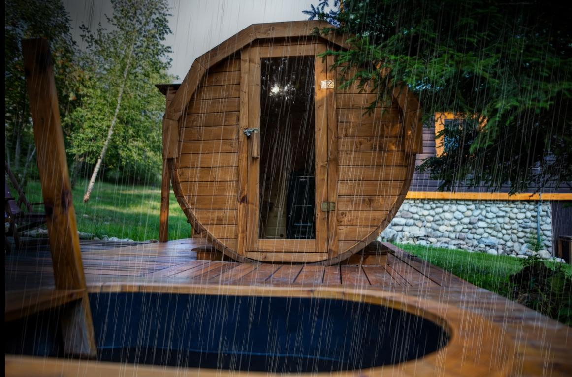 A round barrel sauna sits on a deck next to a hot tub, surrounded by grass and trees, as rain falls heavily, creating a cosy outdoor sauna retreat.