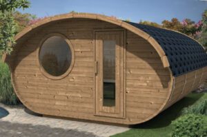 A small, modern wooden cabin with a curved roof, round window, and glass door sits on a stone patio amid lush greenery and vibrant trees, beside the Oval Sauna 4.0m "Hobbit"—ideal for a relaxing hobbit garden retreat.