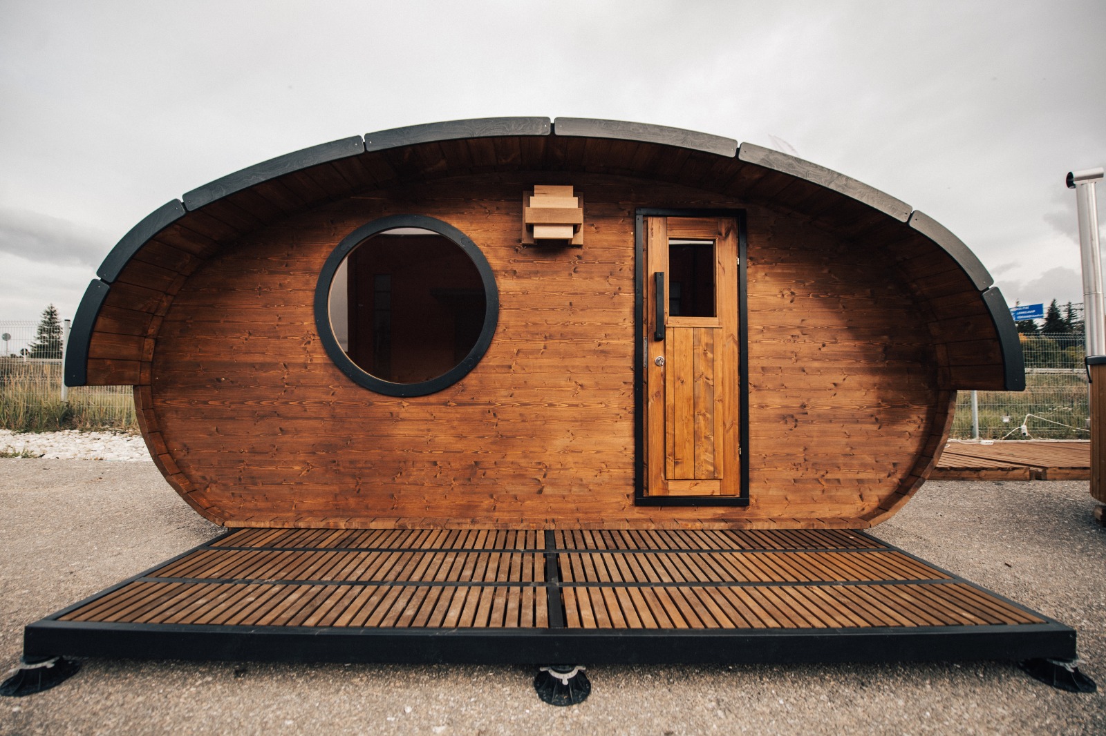 An Oval Sauna 4.0m "Hobbit"—a barrel-shaped wooden cabin with a round window, rectangular door, and wooden deck—stands outdoors in a charming hobbit garden on a cloudy day.